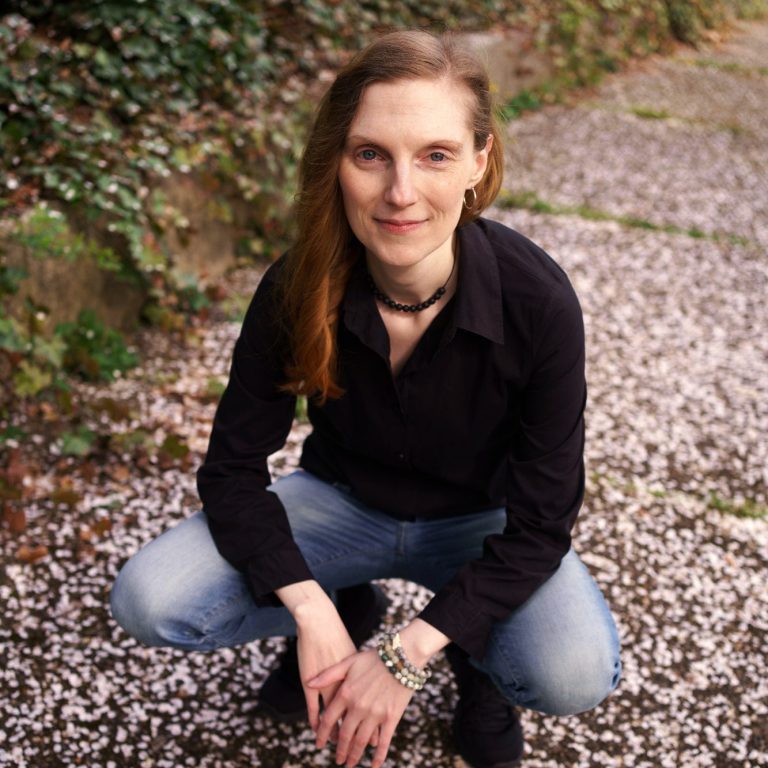 Frau mit rotem Haar in schwarzer Lederjacke sitzt auf einem Stein in der Natur.