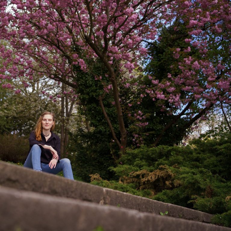 Blick in die Natur Frau mit roten Haaren sitzt am Ufer eines ruhigen Gewässers, umgeben von Bäumen.